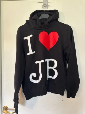 Svart hoodie med I ❤️ JB-tryck storlek S - Svart hoodie med stor vit text 'I ❤️ JB' på framsidan och ett rött hjärta. Hoodien har huva och ribbade muddar vid ärmar och nederkant. Perfekt för dig som gillar statement-plagg och vill sticka ut lite extra. Storlek S.