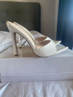 Vita stilettklackade pumps - Säljer ett par vita pumps med öppen tå och spetsig front. Skorna har hög stilettklack och bred rem över foten. Materialet ser ut att vara glansigt, troligen konstläder. Perfekta för dig som vill ha en clean och trendig look.