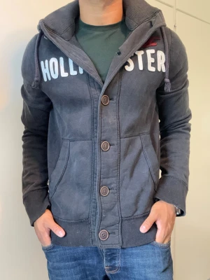Vintage Hollister Cardigan Med Knappar - Väldigt sällsynt och vintage Hollister hoodie med knappar | Tröjan är i bra skick, små defekter förekommer som små hål, se sista bild| Längd ≈ 63cm, Bredd armhåla-amrhåla ≈ 53cm | Modell 175cm, 68kg | Hör av vid ytterligare frågor och priset kan diskuteras vid snabbt köp! |
