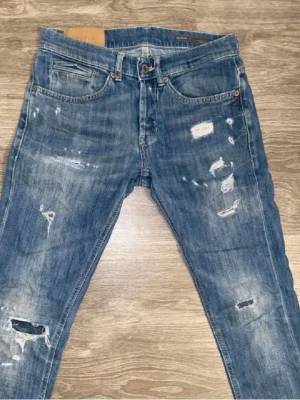 Blå slitna jeans från dondup - Säljer ett par blå jeans från dondup med distressed detaljer och slitningar på både fram- och baksida. Klassisk femficksmodell med knappgylf och raka ben. Jeansen har en cool, avslappnad look och är tillverkade i bomullsdenim.