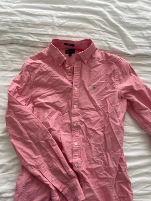 Rosa skjorta från Gant - Klassisk rosa skjorta från Gant med button-down krage och vita knappar. Skjortan har lång ärm och en liten broderad logga på bröstet. Tillverkad i mjuk bomull och passar perfekt till både jeans och chinos.