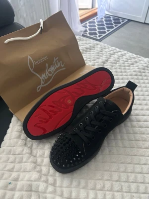 Christian Louboutin  - Säljer ett par svarta sneakers från Christian Louboutin med klassisk röd sula och svarta nitar på tån. Skorna har snörning, är låga och har en lyxig känsla med detaljer i mocka och läder. Perfekta för dig som vill sticka ut med exklusiv stil.