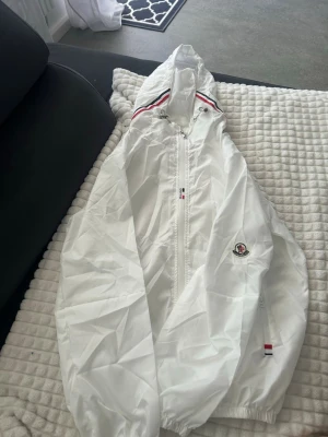 Moncler windbraker - Snygg vit vindjacka från Moncler med huva och dragkedja framtill. Jackan har Moncler-logga på ärmen och detaljer i rött, vitt och blått vid huvan och dragkedjan. Materialet är lätt och glansigt, perfekt för att skydda mot vind.