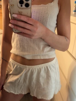 Krämvit pyjamas från lager 157 - Sött tvådelat set från lager 157 i krämvit färg. Setet består av ett ribbat linne med spetskantade axelband och matchande shorts med elastisk midja och vågig kant. Materialet är mjukt och stretchigt, perfekt för att chilla hemma.