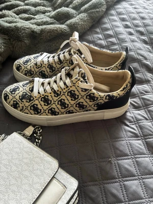 Mönstrade sneakers från Guess - Snygga sneakers från Guess med beige och mörkblått monogrammönster, vita snören och svarta detaljer vid hälen. Guess-logga i guld på baksidan av sulan. Ovandelen är i textil och syntet, sulan är vit och platt.