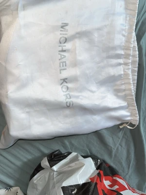 Vit axelväska Michael Kors - Snygg vit axelväska från Michael Kors med silverfärgad kedjerem och diskret logomönster. Väskan har en stilren metallplatta framtill och är tillverkad i tåligt konstläder. Perfekt för dig som vill ha en elegant accessoar.