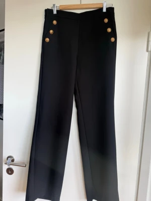 Svarta kostymbyxor med guldknappar - Snygga svarta kostymbyxor med hög midja och raka ben. Fyra dekorativa guldknappar på varje sida framtill ger en cool marininspirerad look. 