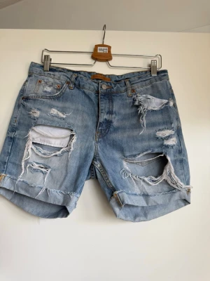Blå  jeansshorts med hål - Säljer ett par ljusblå jeansshorts med rejält slitna detaljer och stora hål framtill. Shortsen har upprullade benslut, klassisk femficksmodell och knappgylf. 