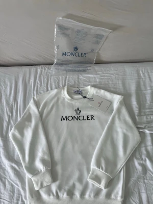 Vit sweatshirt från Moncler - Snygg vit sweatshirt från Moncler med svart logotyptryck på bröstet och Moncler-märke på ärmen. Tröjan har rund halsringning, ribbade muddar och är tillverkad i mjukt material. Perfekt för en clean och stilren look.