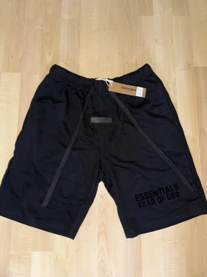 Svarta shorts Essentials Fear of God - Svarta shorts från Essentials Fear of God med långa snören i midjan och tryckt logga på vänster ben. Modellen har elastisk midja och är tillverkad i mjuk bomull, perfekt för en avslappnad streetwear-look.