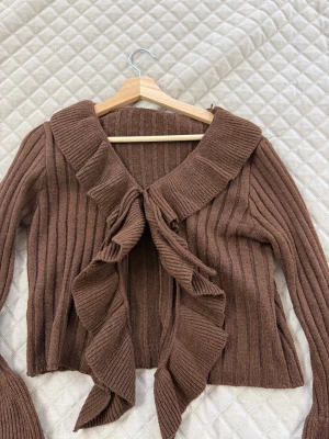 Brun ribbstickad cardigan med volang - Säljer en brun ribbstickad cardigan med stora volangdetaljer framtill och vid ärmsluten. Tröjan har en bred krage och är öppen framtill utan knäppning. Perfekt för dig som gillar en unik och trendig look.
