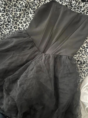 Svart axelbandslös tyllklänning - Snygg svart axelbandslös klänning med figurnära överdel och voluminös kjol i tyll. Perfekt för dig som vill ha en klassisk men ändå trendig look. Kjolen har flera lager för extra volym och överdelen är slät och enkel.