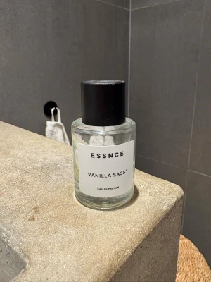 ESSNCE Vanilla Sass Eau de Parfum - Fräsch parfym från ESSNCE med doft av vanilj. Flaskan är cylindrisk i genomskinligt glas med svart lock och enkel vit etikett. Perfekt för dig som gillar söta och mjuka dofter med en modern touch.