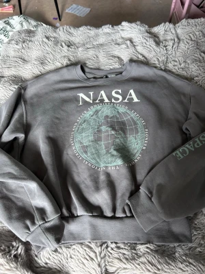 Grå NASA sweatshirt med jordglob - Cool grå sweatshirt med stort NASA-tryck och en jordglob på bröstet. Tröjan har ribbade muddar vid ärmslut och nederkant samt ett SPACE-tryck på ena ärmen. Perfekt för dig som gillar rymden och vill ha en avslappnad stil.