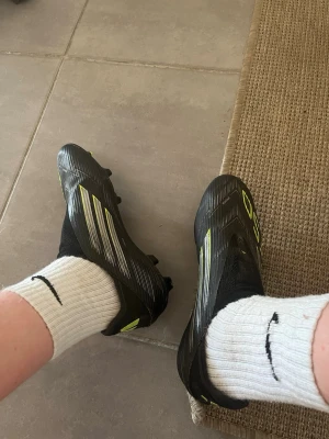 Adidas svarta fotbollsskor med neon - Säljer ett par svarta Adidas fotbollsskor med neon-gula detaljer och vita ränder. Skorna har en modern, strömlinjeformad design och är tillverkade i syntetmaterial med fasta dobbar för spel på gräs. Perfekta för dig som vill ha snabba och lätta skor på planen.