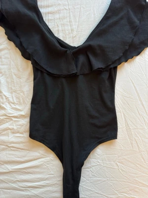 Svart body med volang - Snygg svart body med djup v-ringning och bred volang runt axlarna. Tillverkad i mjukt stretchigt tyg som sitter skönt mot kroppen. Perfekt för dig som vill ha en enkel men ändå trendig look.