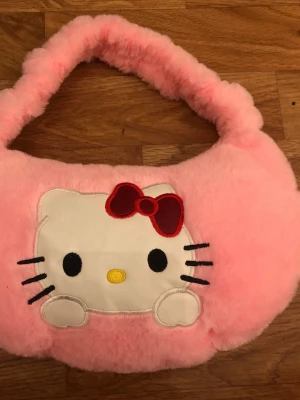 Plyschig rosa Hello Kitty handväska - Supergullig rosa handväska i mjuk plysch med Hello Kitty-motiv framtill. Väskan har ett fluffigt handtag och en broderad Hello Kitty med röd rosett. Perfekt för dig som älskar söta accessoarer och vill sticka ut med en unik väska.