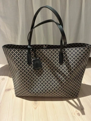  shoppingväska från By Malene Birger i mycket bra skick! - Snygg shoppingväska från By Malene Birger i svart och beige med klassiskt grafiskt mönster. Väskan har svarta handtag i konstläder och en rymlig insida med ljusrosa foder och innerfack med dragkedja. Perfekt för dig som vill ha plats för allt. Mått höjd 28 cm botten 14 x 37 cm.
