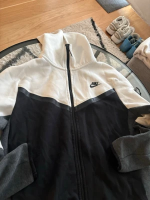 Nike tech vintage hoodie  - Nike hoodie med dragkedja i svart och vitt med grå detaljer på ärmarna. Klassisk logga på bröstet och huva. Tillverkad i mjukt material, perfekt för en avslappnad streetwear-look.
