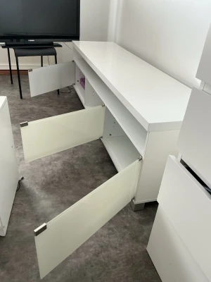 Vit tv-bänk med luckor - Säljer en stilren vit tv-bänk med fyra stora luckor som öppnas utåt. Bänken har en lång, rektangulär form och är tillverkad i trä med en blank finish. Perfekt för förvaring av elektronik, spel eller andra prylar i vardagsrummet.