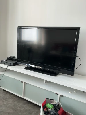 Svart platt-TV från Sony - Säljer en svart platt-TV från Sony med bred ram och stabil fot. TV:n har flera anslutningsmöjligheter på sidan, inklusive HDMI och komponentingångar. Perfekt för gaming eller filmkvällar. Klassisk design som passar i de flesta vardagsrum.