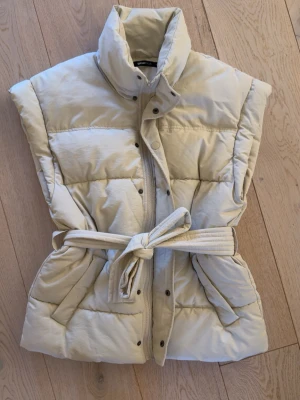Beige dunväst med bälte - Stilren beige dunväst med hög krage och knytbart bälte i midjan. Västen har tryckknappar och dragkedja framtill samt två snedställda fickor. Perfekt för lager-på-lager och ger en trendig siluett. Materialet är quiltat och vadderat för extra värme.