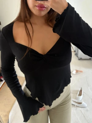 Svart långärmad topp med twist - Trendig svart långärmad topp med twistad detalj framtill och v-ringad urringning. Toppen har vågig kant nertill och vida ärmslut för en cool look. Perfekt att styla med både jeans och kjol.