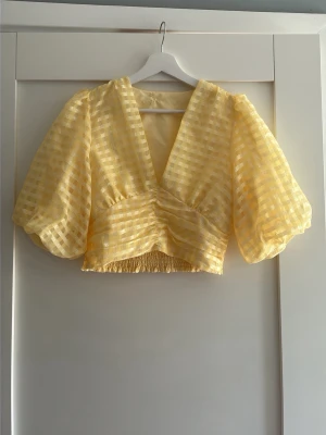 Gul croppad blus med puffärmar - Säljer en ljusgul croppad blus med v-ringning och stora puffärmar. Blusen har ett rutigt mönster i transparent material och smockad midja. Perfekt för dig som vill ha en trendig och luftig topp till sommaren.