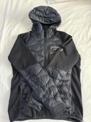 Svart pufferjacka från Peak Performance - Snygg svart pufferjacka från Peak Performance med huva och dragkedja framtill. Jackan har quiltade partier på bröst, rygg och ärmar samt släta svarta sidopaneler. Perfekt för kalla dagar och riktigt skön passform. Logga på bröstet och ärmen.