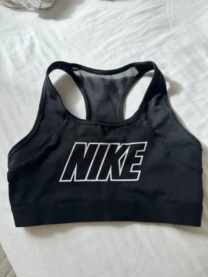 Svart sport-bh från Nike - Svart sport-bh från Nike med stort vitt logotryck framtill. Bh:n har brottarrygg och är tillverkad i ett stretchigt, fukttransporterande material som passar perfekt för träning. Snygg och funktionell design med bred resår nedtill för extra stöd.