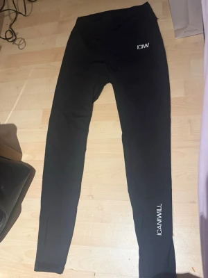 Svarta leggings från ICANIWILL - Säljer ett par svarta leggings från ICANIWILL med logga på höften och längs benet. De har hög midja och är tillverkade i ett stretchigt material som sitter tight. Perfekta för träning eller chill. Snygg och enkel design.