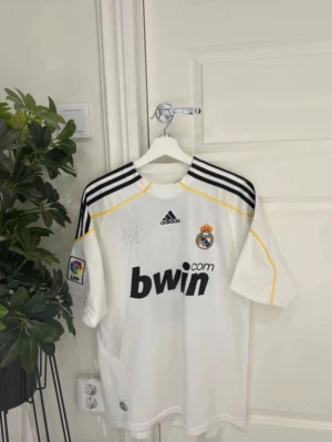 Real Madrid Ronaldo 9 matchtröja - Säljer en vit Real Madrid matchtröja från Adidas med svarta detaljer och gula inslag. Tröjan har klubbmärke, Adidas-logga och Bwin som sponsor framtill. På ryggen står det Ronaldo och nummer 9. Korta ärmar och broderade detaljer. Perfekt för fotbollsfans!