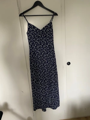 Marinblå blommig långklänning - Snygg marinblå långklänning med vita små blommor, slits nere vid benet och smala axelband. Klänningen har v-ringad hals och figurnära passform. Perfekt för varma dagar och somriga tillfällen.