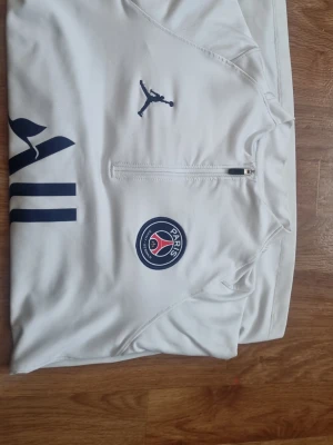 Vit PSG träningströja Jordan - Snygg vit träningströja från PSG x Jordan med halv dragkedja framtill. Tröjan har Paris Saint-Germain klubbmärke på bröstet och mörkblå detaljer, inklusive Jordan-loggan och ränder på sidan. Tillverkad i ett mjukt och stretchigt funktionsmaterial.