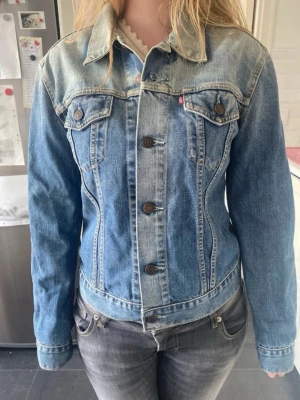 Levi's klassisk blå jeansjacka - Snygg blå jeansjacka från Levi's med klassisk passform och markerade sömmar. Jackan har två bröstfickor med knappar, metallknappar framtill och en liten röd Levi's-tagg. Perfekt till alla dina outfits!
