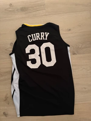 Svart basketlinne Curry #30 - Snyggt svart basketlinne med vita detaljer och gult runt kragen. På ryggen står det 'Curry' och nummer 30, framtill står det 'The Town' med ett trädmotiv. Perfekt för basket eller streetwear.