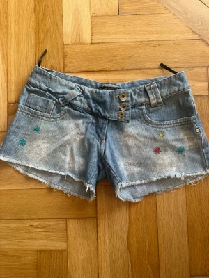 Vintage jeansshorts  - Snygga jeansshorts i ljus denim med färgglada broderade stjärnor framtill och baktill köpta i Grekland. Modellen har låg midja, råa fransade kanter och dekorativa knappar som ger en härlig Y2K- och sommarkänsla.
