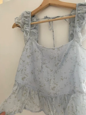 Ljusblå blommig linnetopp med volang - Supersöt ljusblå linnetopp med små blommor, volangdetaljer på axelbanden och nedtill samt knyt i nacken. Toppen har smock i ryggen för extra komfort och en luftig, somrig känsla. Perfekt för dig som gillar romantisk och feminin stil.
