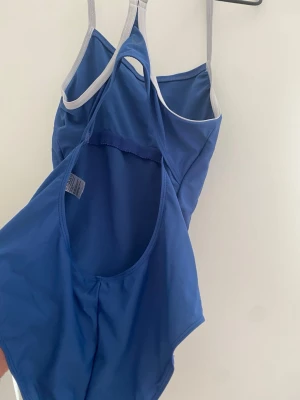 Blå baddräkt med vita band - En stilren blå baddräkt med tunna vita axelband som korsas i ryggen. Klassisk modell med öppen rygg och mjukt, stretchigt material som passar perfekt för simning eller strandhäng.