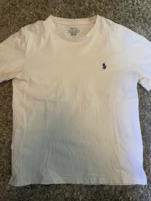 Vit t-shirt från Polo Ralph Lauren - Klassisk vit t-shirt från Polo Ralph Lauren med rund halsringning och korta ärmar. T-shirten har den ikoniska lilla marinblå logotypen broderad på bröstet. Tillverkad i mjuk bomull för en skön känsla och enkel stil.
