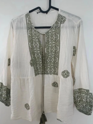 Vit boho-blus med brodyr från Zara - Luftig vit blus från Zara med gröna broderade detaljer och knytband med tofsar vid halsen. Blusen har lång ärm och ett avslappnat, bohemiskt snitt. Tillverkad i ett tunt, strukturerat tyg som ger en skön känsla.