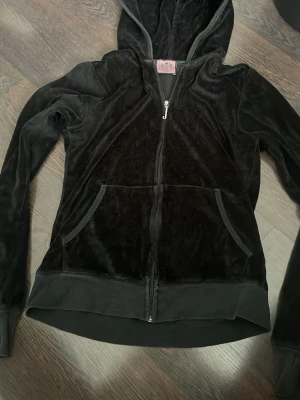 Svart velourhoodie från Juicy Couture - Säljer en svart velourhoodie från Juicy Couture med dragkedja framtill och klassisk huva. Hoodien har kängurufickor och ribbade muddar vid ärmslut och nederkant. Perfekt för en avslappnad och trendig look.