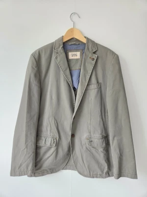 Grå kavaj från Camel Active - Stilren grå kavaj från Camel Active med klassisk passform och tvåknäppning. Kavajen har tre framfickor, slits bak och är tillverkad i ett mjukt bomullstyg med diskret struktur. Perfekt för en avslappnad men ändå uppklädd look.