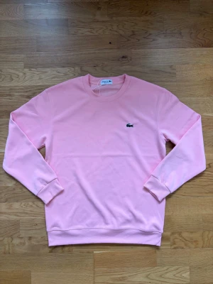 Ljusrosa sweatshirt från Lacoste - Säljer en ljusrosa sweatshirt från Lacoste med klassisk rund halsringning och det ikoniska krokodilmärket broderat på bröstet. Tröjan har ribbade muddar vid ärmslut och nederkant. Perfekt för en avslappnad och stilren look.