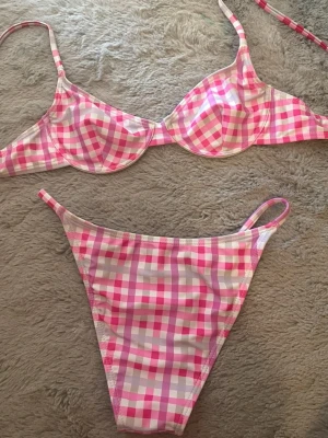 Rutig bikini i rosa och lila toner - Söt bikini med rutigt mönster i rosa, lila och vitt. Överdelen har bygel och smala axelband, underdelen har högt skurna ben och tunna sidoband. Perfekt för strandhäng eller poolparty! Materialet känns mjukt och följsamt.