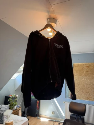 Brun FSBN hoodie - Svart oversized hoodie från FSBN med dragkedja framtill och tryck på bröstet. Hoodien har huva och långa armar, perfekt för en avslappnad streetwear-look. Materialet känns mjukt och bekvämt, och modellen är loose fit.