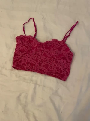 Rosa spetsig bralette med smala band - Säljer en snygg rosa bralette i spets med tunna axelband och blommigt mönster. Braletten har en kort, croppad modell och är tillverkad i stretchigt spetsmaterial som känns mjukt mot huden. Perfekt att bära under en skjorta eller som den är.