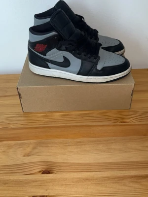 Air Jordan 1 Mid svart/grå/röd - Snygga Air Jordan 1 Mid sneakers i svart och grått med röda detaljer på sidan. Klassisk siluett med svart Swoosh och perforerad tå. Skorna har högt skaft och är tillverkade i läder och syntetmaterial. Perfekta för dig som gillar streetwear och ikoniska sneakers.