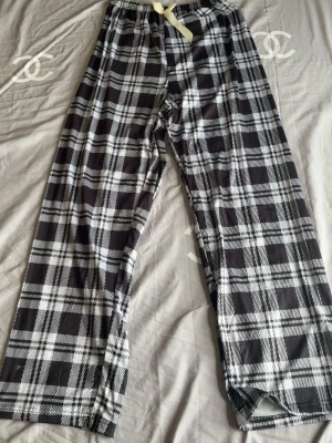 Rutiga pyjamasbyxor med snöre - Säljer ett par klassiska pyjamasbyxor i svartvitt rutigt mönster. Byxorna har ett bekvämt midjeband med vitt snöre för justering och är tillverkade i ett mjukt, lätt material. Perfekta för chillkvällar hemma.
