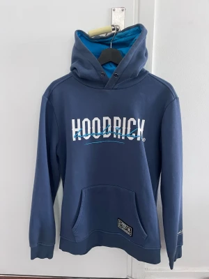 Blå hoodie från Hoodrich med tryck - Snygg blå hoodie från Hoodrich med vit och blå logga framtill. Hoodien har en stor huva med ljusblå insida, känguruficka och ribbade muddar. Tillverkad i mjukt bomullsmaterial, perfekt för en avslappnad streetwear-look.
